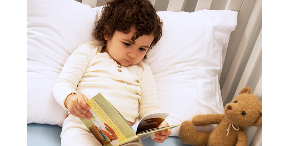 Toddler laying in bed reading a book with teddy bear next to her.