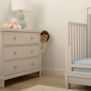 Smiling toddler hiding next to dresser while poking head out. 