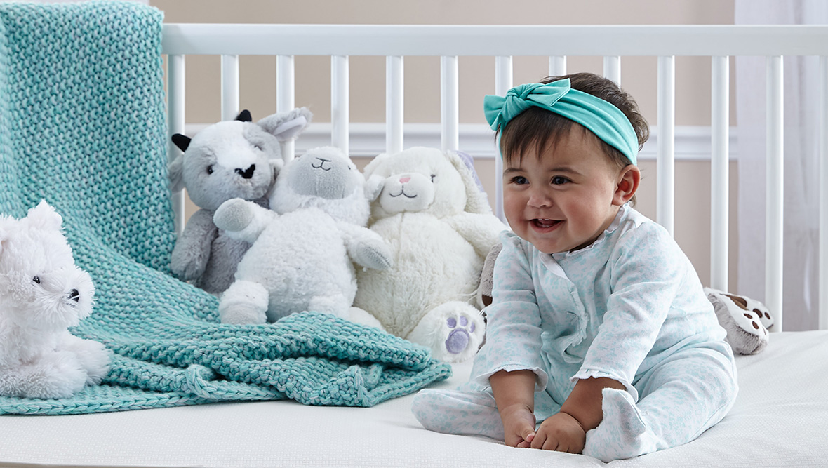 Smiling baby sitting in her crib mattress with a blanket and stuffed animals. 