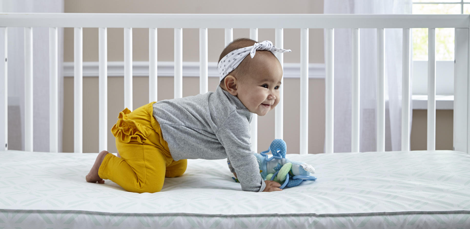 Smiling baby crawling inside crib mattress with toy in her hand. 